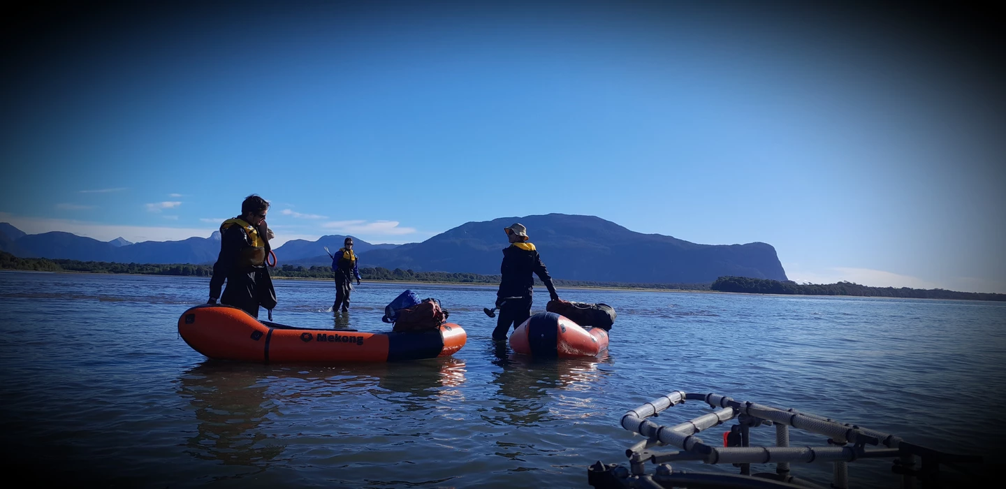 Vier Personen mit orangefarbenen Packrafts auf ruhigem Gewässer, Berglandschaft im Hintergrund