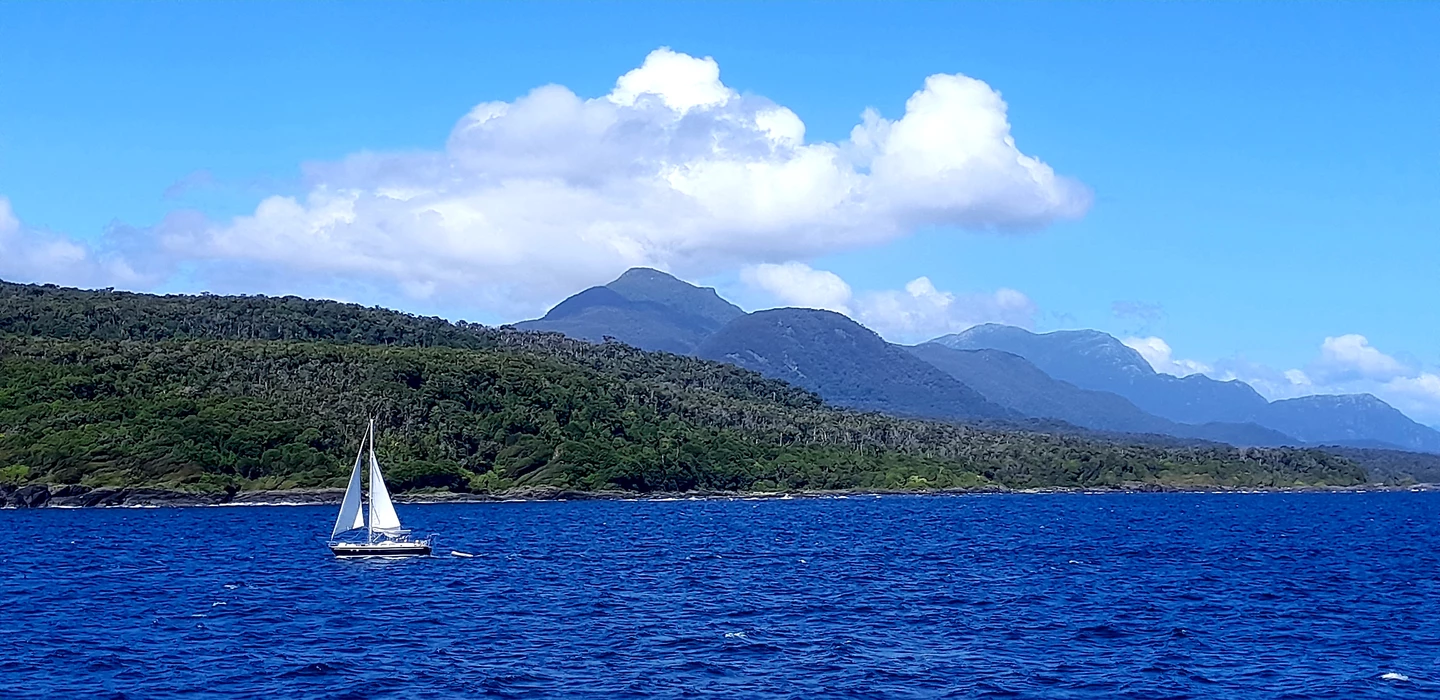 Blauer See mit weißem Segelboot, im Hintergrund dicht bewaldete Berge unter wolkigem Himmel