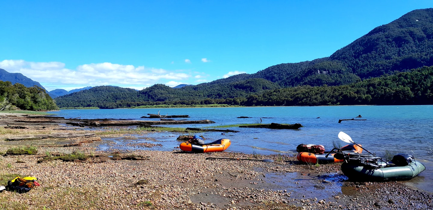 Zwei orange-graue Packrafts an felsigem Seeufer, im Hintergrund bewaldete Berge und blauer Himmel