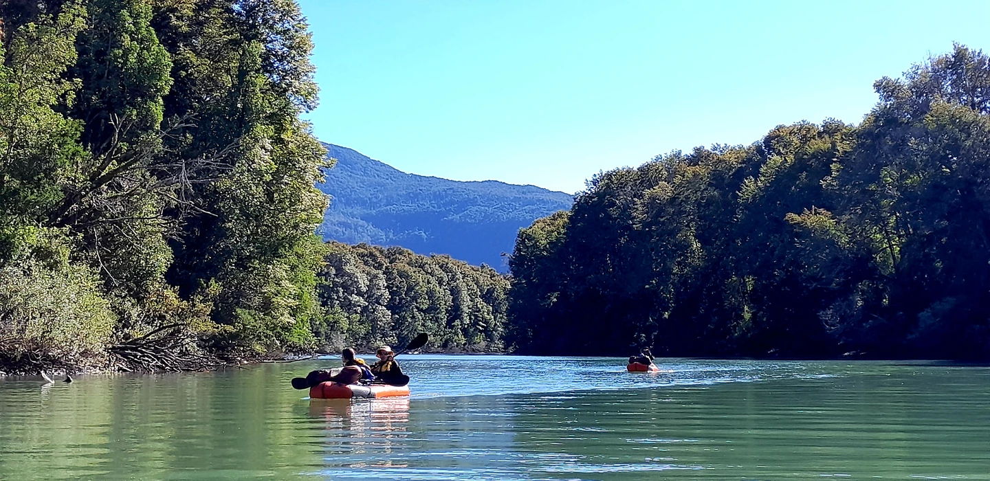 Zwei Kajaker paddeln auf grün-blauem Fluss, umgeben von dichten Wäldern und Bergen im Hintergrund
