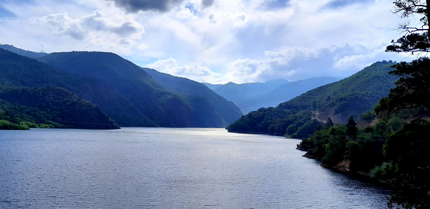 Weite Landschaftsaufnahme eines spiegelglatten Sees zwischen bewaldeten Berghängen unter wolkigem Himmel