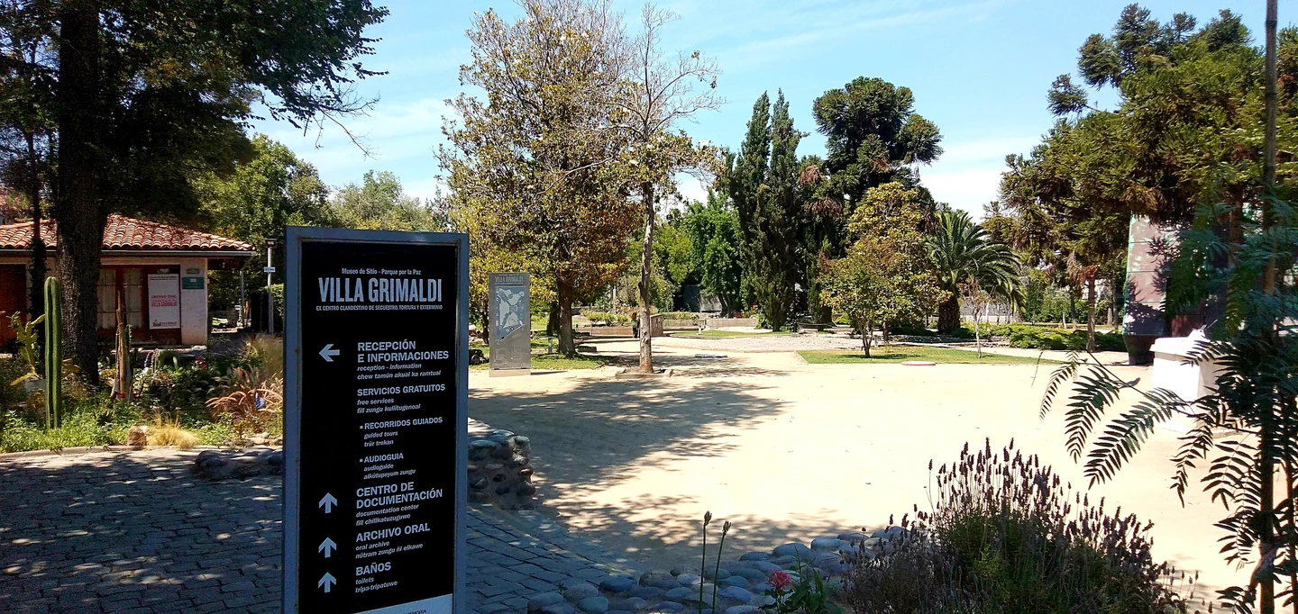 Infotafel von Villa Grimaldi mit Parkanlage, Bäumen und Wegweisern im Hintergrund