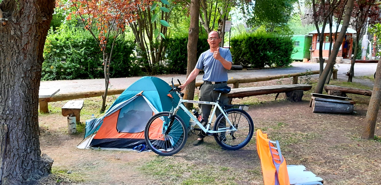 Blau-orangefarbenes Zelt auf Campingplatz, daneben weißes Mountainbike und eine Person im grauen T-Shirt