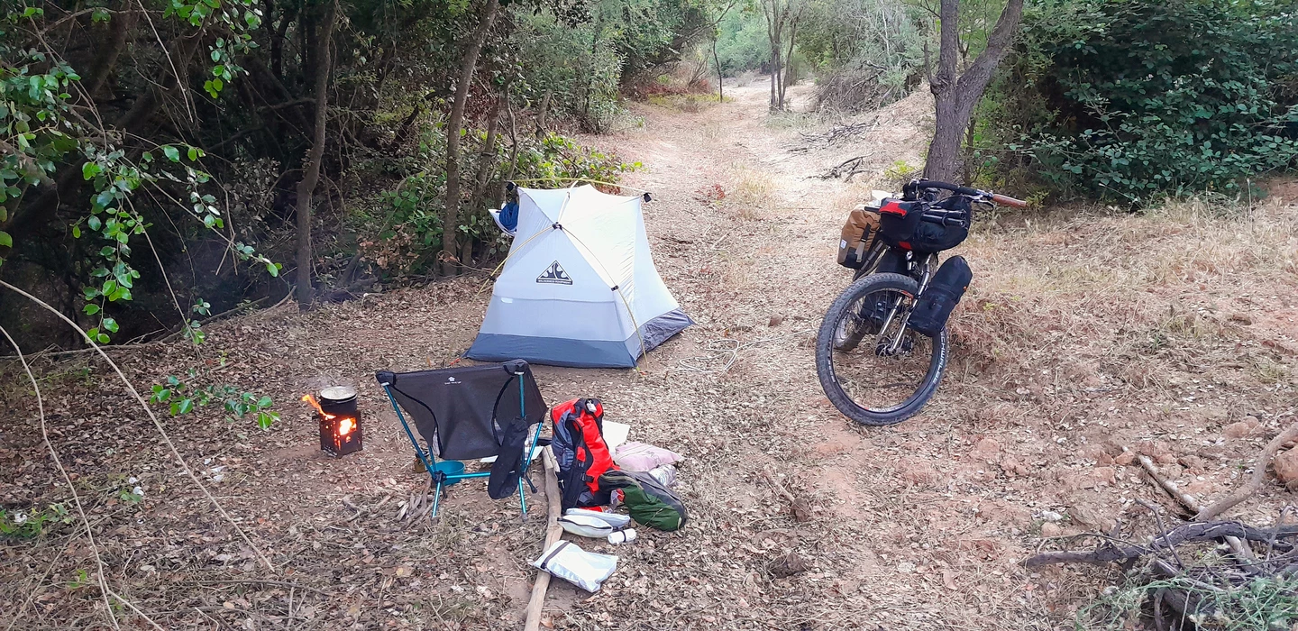 Weißes Zelt, Fahrrad und Campingausrüstung auf einem schmalen Waldweg mit Feuerstelle