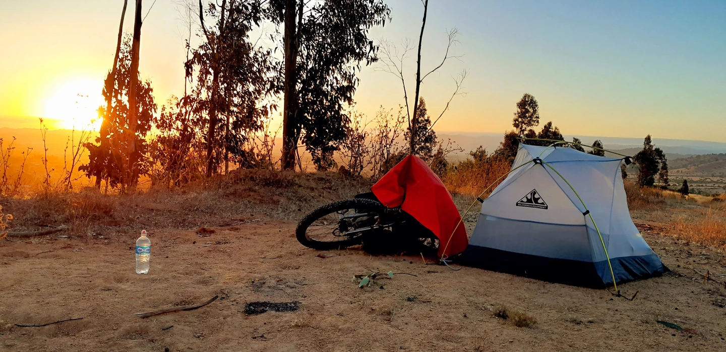 Weißes Zelt und rotes Gepäck neben einem Fahrrad im trockenen Gelände bei Sonnenaufgang, Wasserflasche im Vordergrund