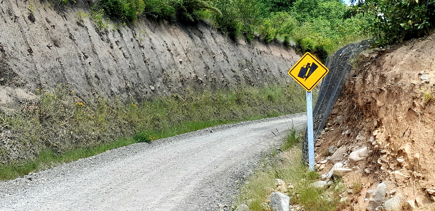 Unbefestigte Schotter-Bergstraße mit gelbem Warnschild für steile Gefälle, links und rechts von Felswänden und Vegetation gesäumt