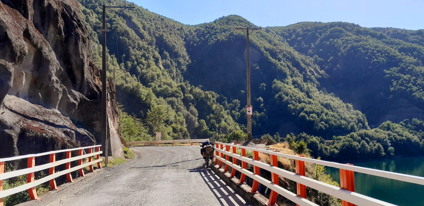 Schmale Brücke mit rot-weißen Geländern über einen grünen See, umgeben von bewaldeten Berghängen und Felswänden