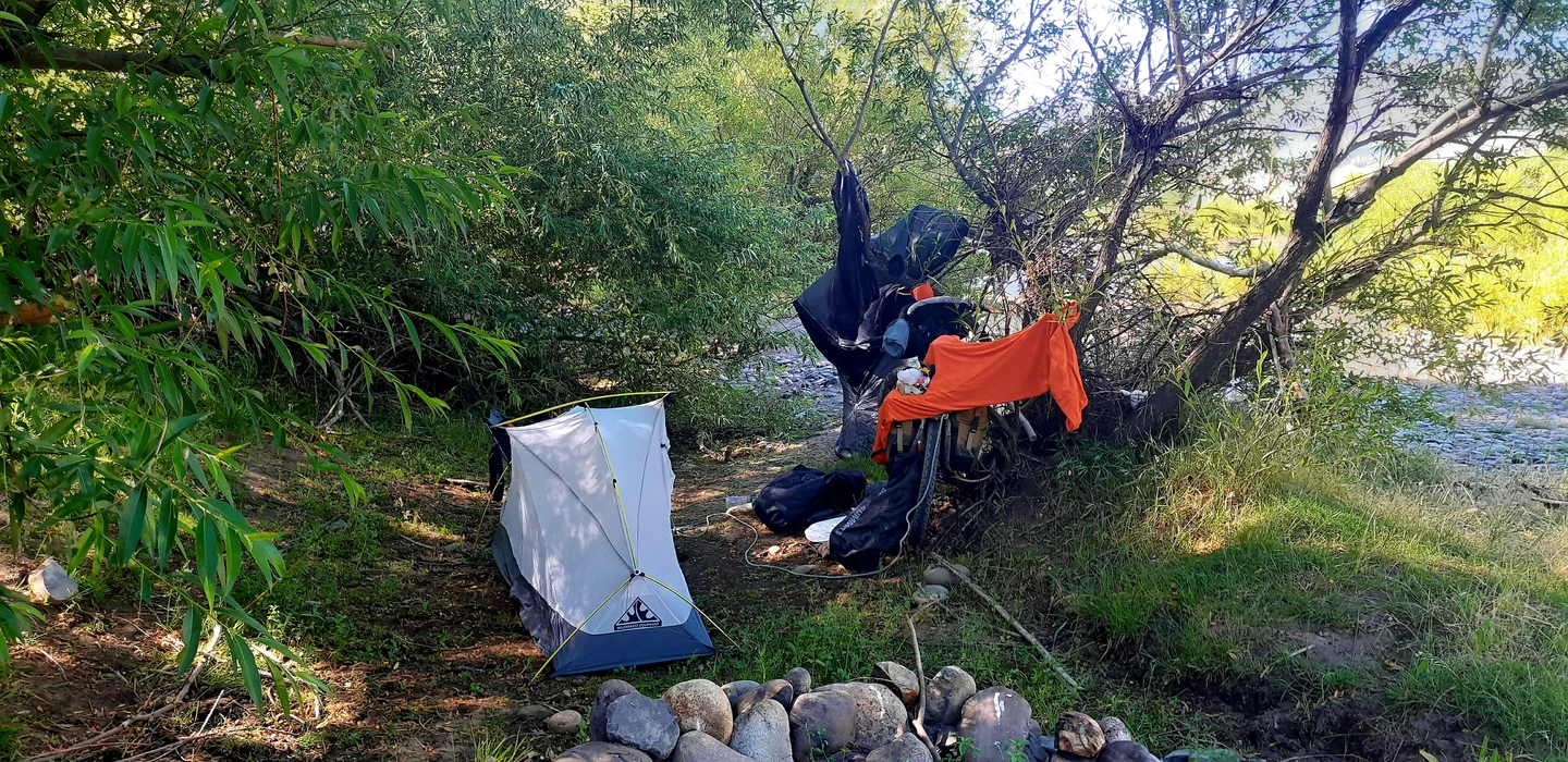Small white tent set up near rocky stream, orange clothing hanging on tree branch, surrounded by green foliage