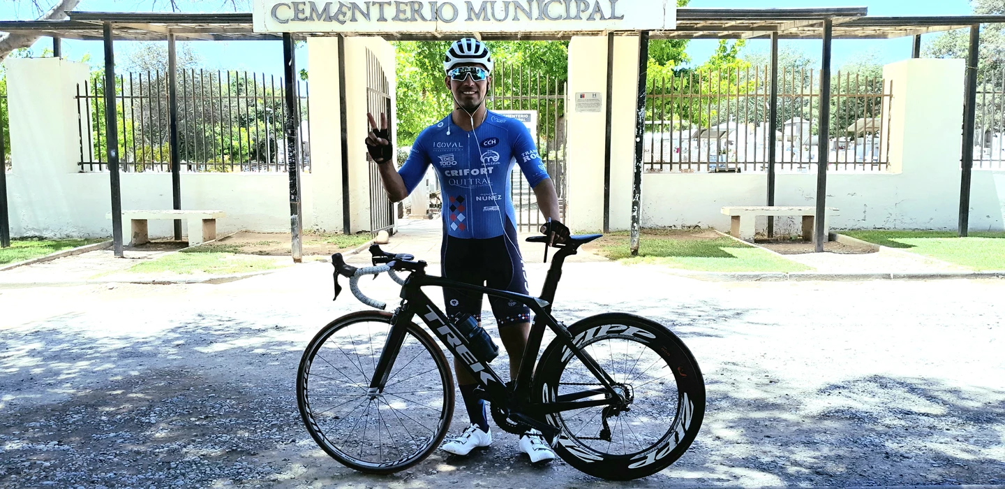 Radfahrer in blauer Sportbekleidung mit Rennrad vor Eingang des Cementerio Municipal, Zaun und Bäume im Hintergrund