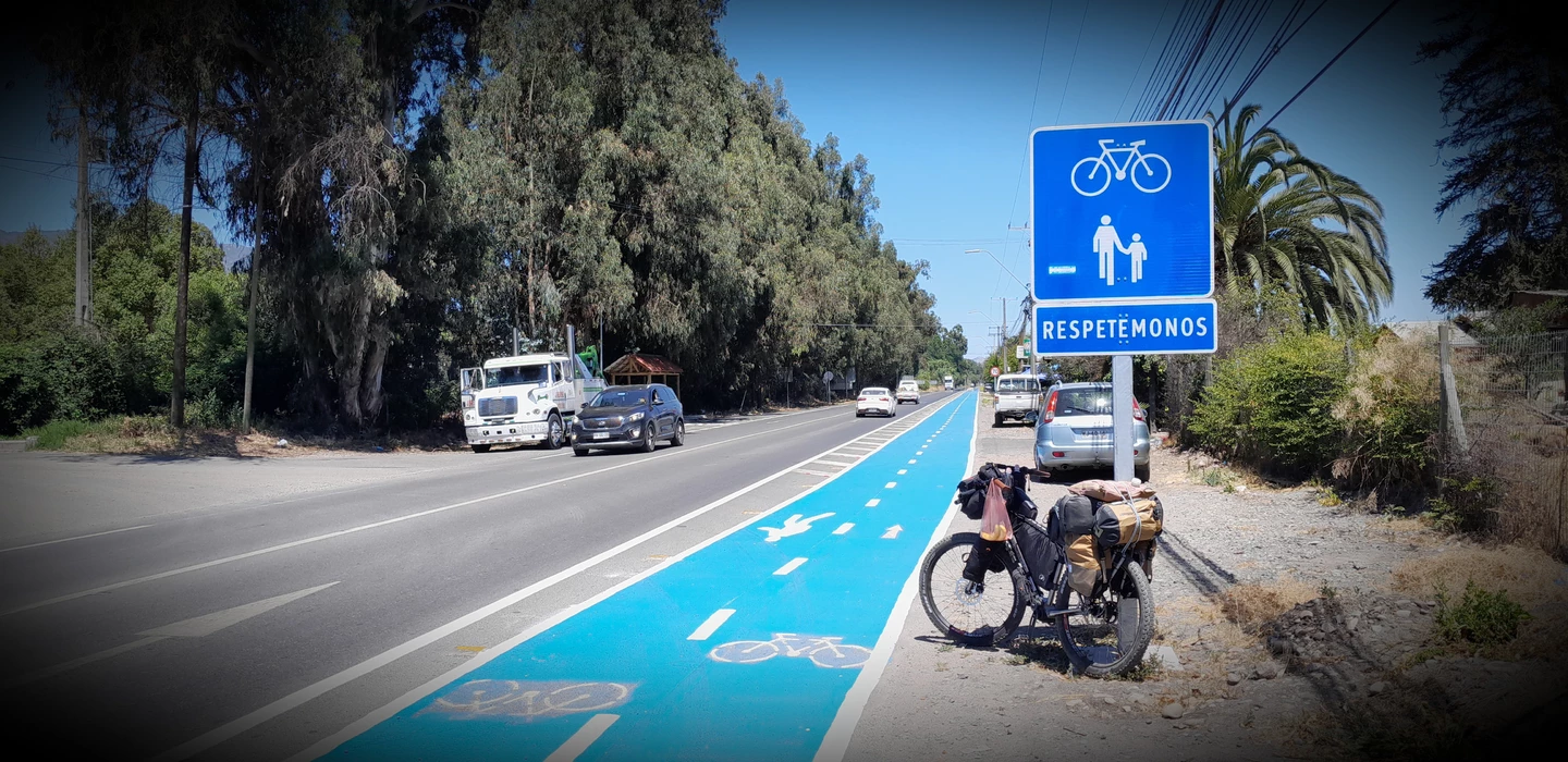 Blau markierter Radweg an Landstraße mit Verkehrsschild 'Respetémonos', Lastwagen und Fahrrad mit Gepäck im Hintergrund