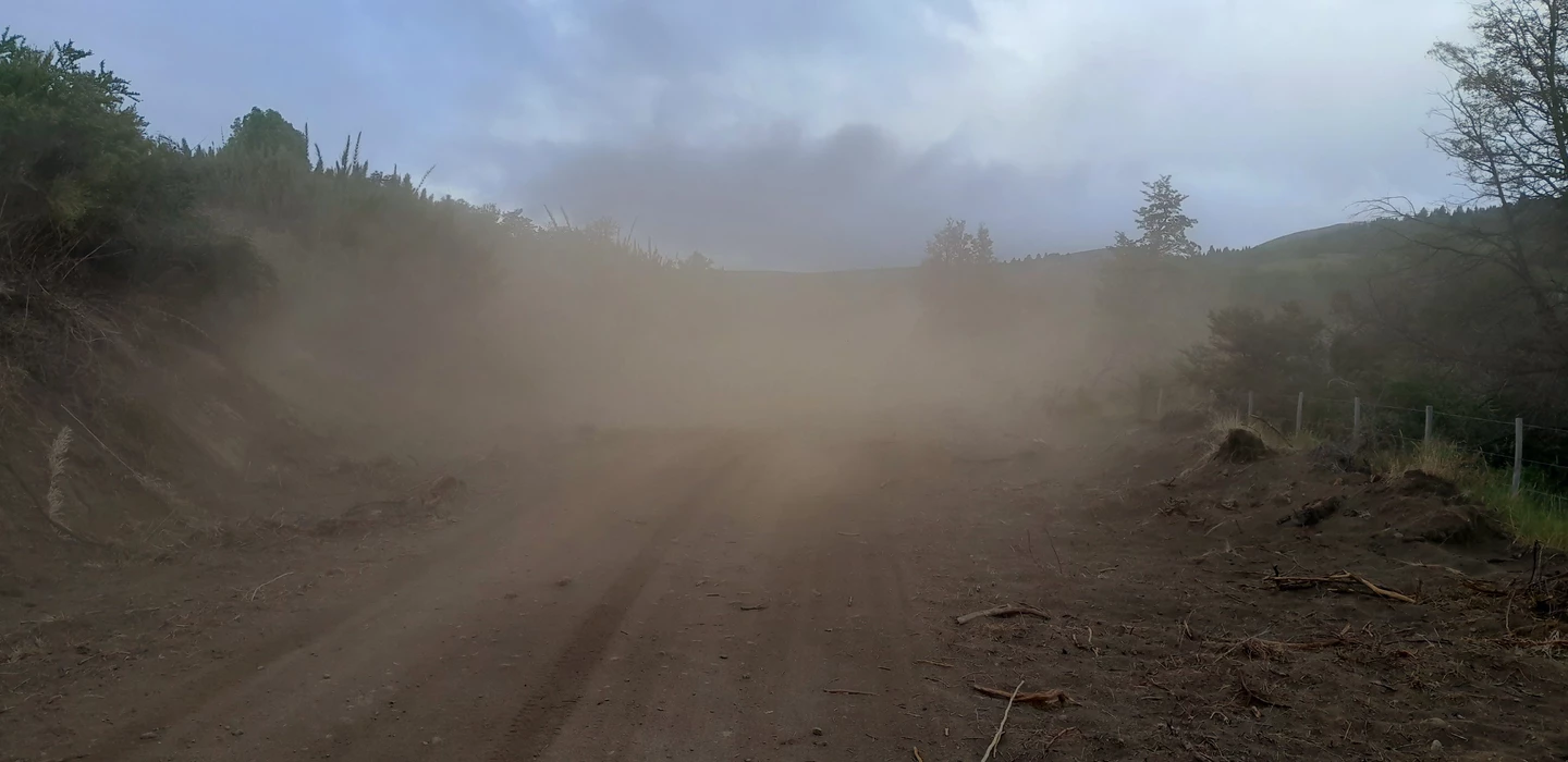 Unbefestigter Feldweg mit dichter Staubwolke, umgeben von Büschen und Zaun, Berghänge im Hintergrund