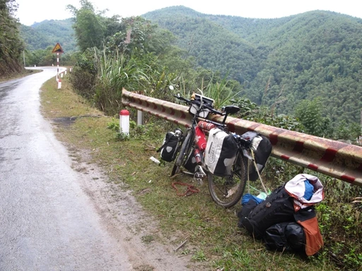 Fahrrad mit Gepäcktaschen an Leitplanke, nasse Bergstraße mit Blick auf dichten Laubwald im Hintergrund