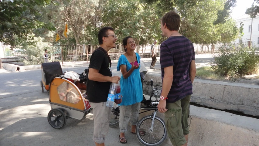 Drei Menschen mit Fahrrädern und Anhänger unterhalten sich auf sonniger Straße mit Bäumen und Zaun
