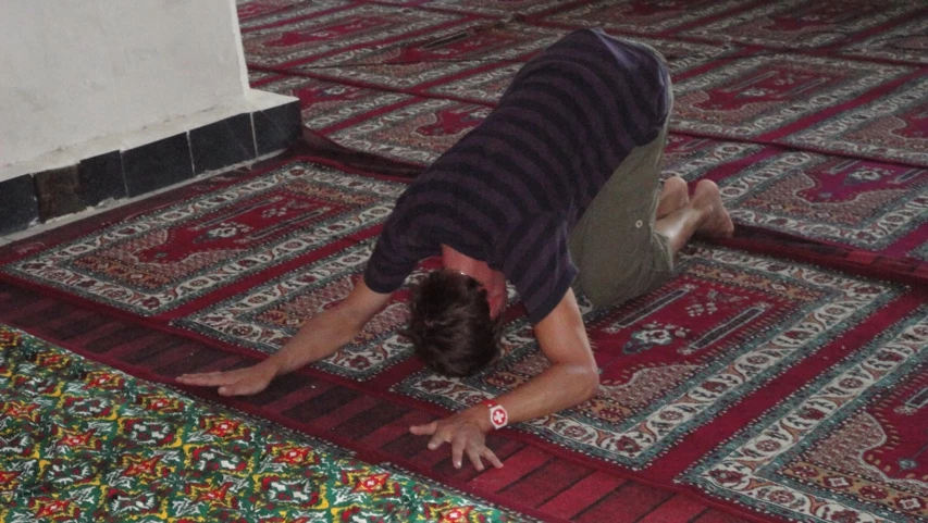 Person in gestreifter Kleidung kniet auf bunten Teppichen und betet in einer Moschee