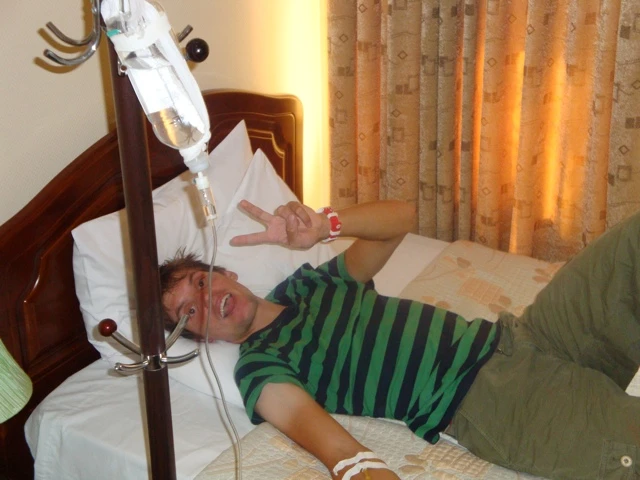 Person in grün-schwarzem Shirt liegt auf Krankenhausbett mit angehängter Infusion und Tropfstativ