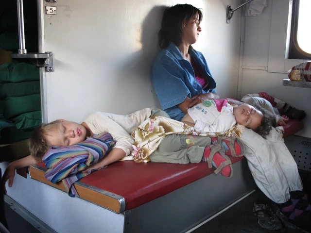 Zwei Kinder schlafen auf roten Sitzen, daneben eine Frau im blauen Kittel, Fenster und Gepäckablage sichtbar