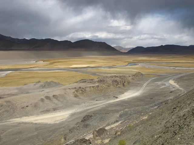 Karges Hochtal mit Schmelzwasserrinnen, Schotterweg und dunklen Bergen unter bewölktem Himmel