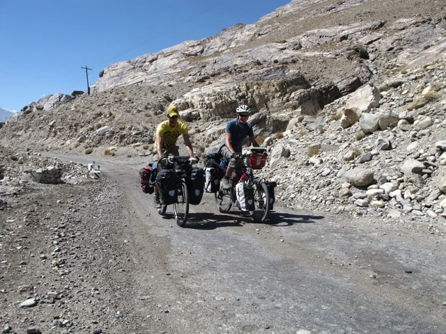 Zwei Radfahrer mit beladenen Fahrrädern auf unbefestigter Bergstraße, Felsenhang im Hintergrund, blauer Himmel