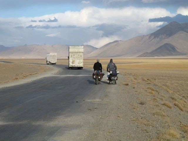 Zwei Radfahrer auf einsamer Straße, im Hintergrund Lastwagen und weite Berglandschaft mit braunem Grasland