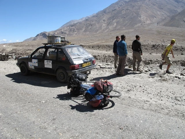 Beschädigte Straßenfahrzeug und Fahrrad in steiniger Berglandschaft mit vier Personen, staubige Geröllpiste mit Bergpanorama im Hintergrund