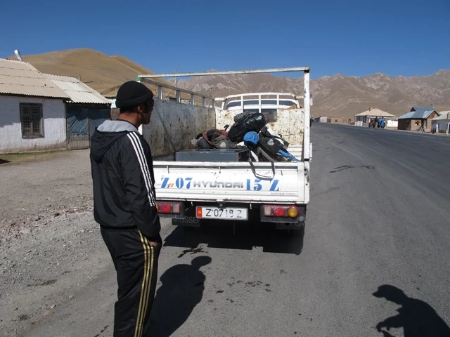 Weißer Hyundai-Transporter mit blauem Motorrad auf Ladefläche, parkend an Bergstraße mit karger Bergkulisse und einfachen Gebäuden
