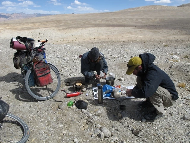 Zwei Radfahrer kochen an einem kargen, steinigen Ort mit Fahrrädern und Campingausrüstung, Berglandschaft im Hintergrund