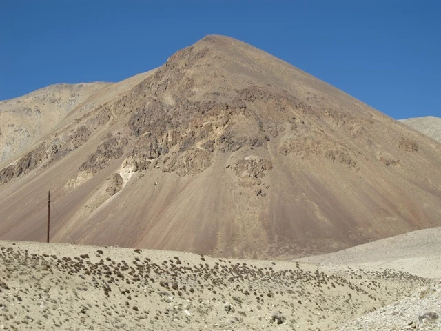 Steile, bräunliche Bergkette mit felsiger Oberfläche und spärlicher Vegetation, vor blauem Himmel und steiniger Ebene