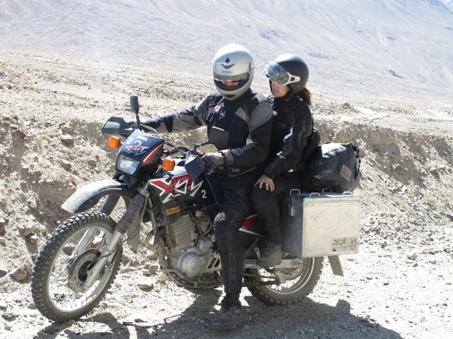 Motorrad mit zwei Fahrern, beladen mit Gepäck, auf steinigem Weg vor felsiger Bergkulisse, Fahrer in schwarzer Motorradausrüstung