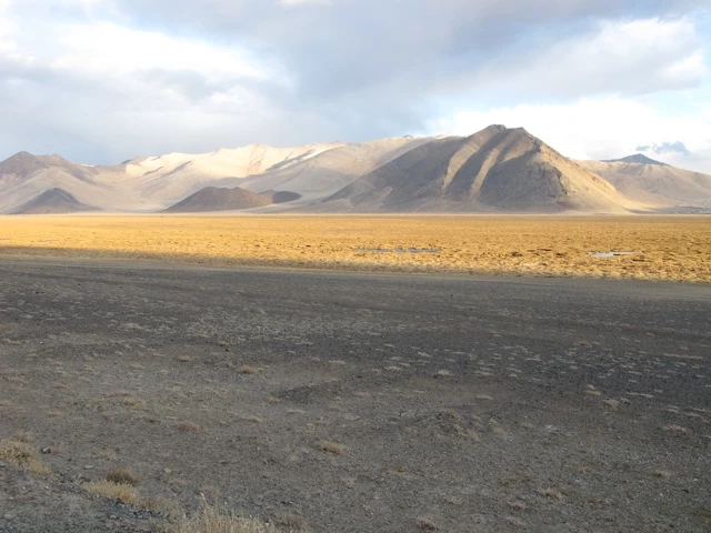 Karges Hochland mit grauen Felsvorsprüngen, gelblich-brauner Ebene und sanft gewellten Bergen im Hintergrund unter bewölktem Himmel