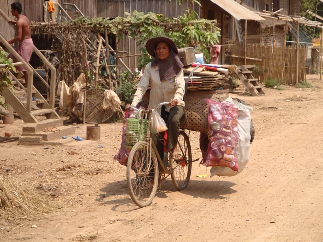 Frau mit Strohhut transportiert Waren auf Fahrrad durch staubige Dorfstraße mit Holzhütten