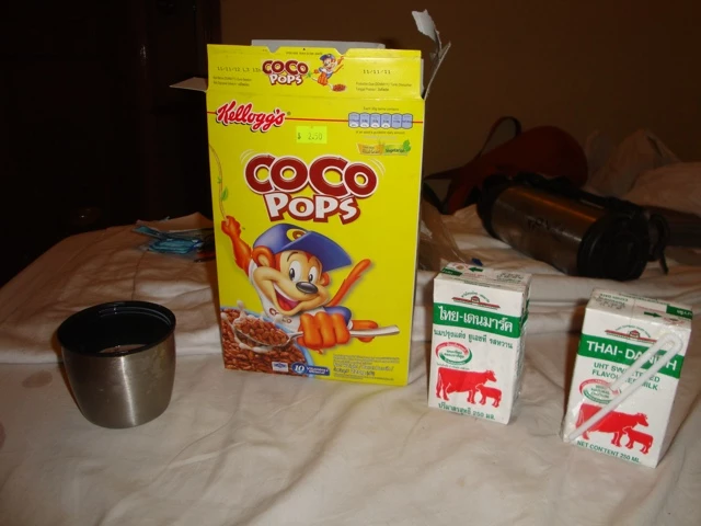 Gelbe Kellogg's Coco Pops Cerealienbox neben zwei weißen Milchkartons und einem Metallbecher auf hellem Untergrund