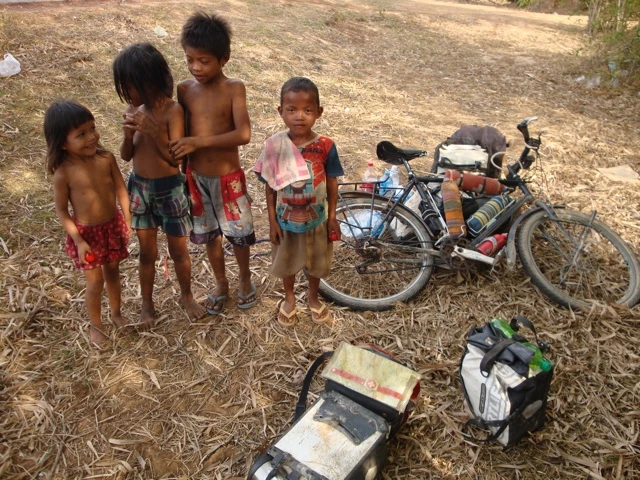 Vier kleine Kinder stehen neben einem Fahrrad auf einer trockenen, strohigen Fläche mit Gepäck und Ausrüstung