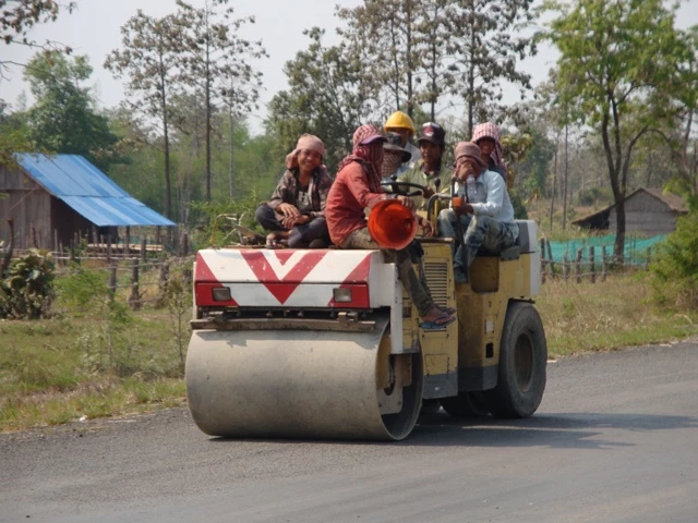 Gelbe Straßenwalze mit mehreren Arbeitern in roten Warnwesten auf einer asphaltierten Landstraße, umgeben von Bäumen und Hütten