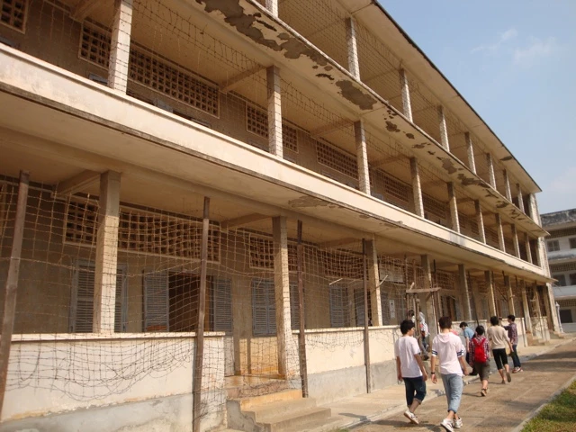 Mehrstöckiges graues Gebäude mit Gitternetzen an Fenstern und Balkonen, Gruppe von Menschen zu Fuß im Vordergrund