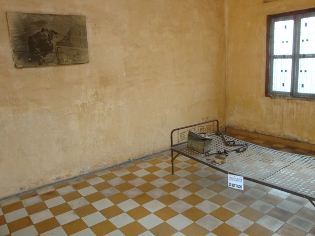 Karges Gefängnis-Zimmer mit gelblicher Wand, kariertem Boden und Metallbett mit Decke, Wandbild und Fenster