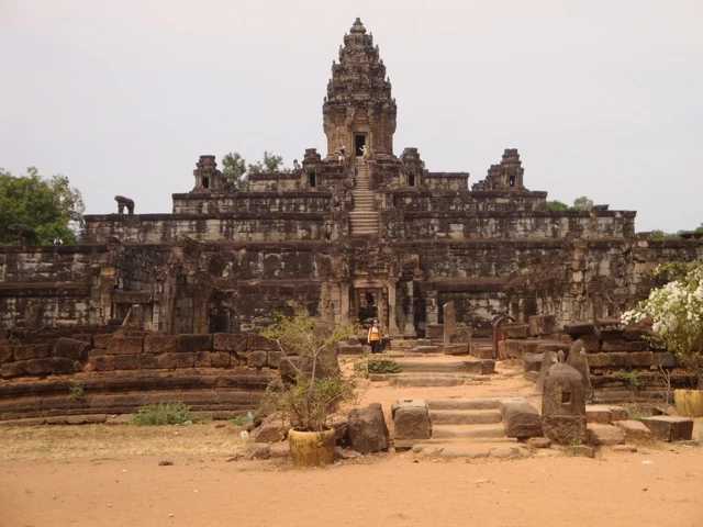 Steinerner Khmer-Tempel mit mehrstufiger Pyramidenstruktur, rötlich-braune Steinblöcke, zentrale Treppe, eine Person in orangefarbener Kleidung