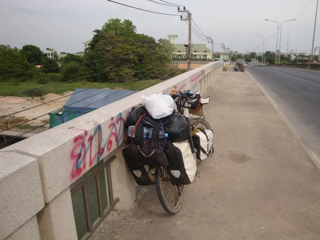 Schwer bepacktes Fahrrad mit Packtaschen an Betonbrücke, Landstraße im Hintergrund, Bäume und Strommasten