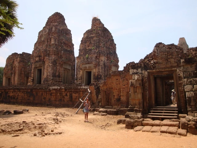 Verfallene Sandsteintempel mit hohen Türmen in rötlich-brauner Farbe, Besucher mit Rucksack im Vordergrund auf sandigem Boden