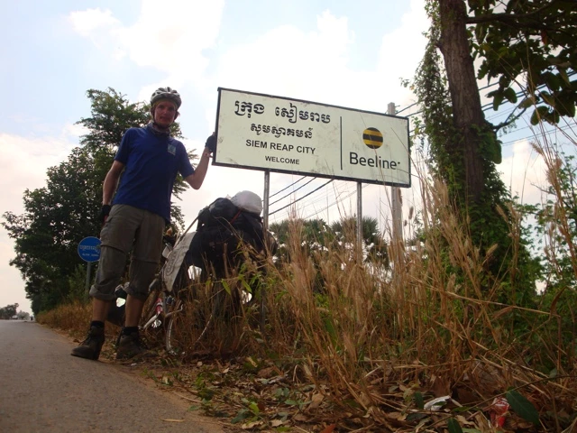 Radfahrer steht neben Willkommensschild von Siem Reap City mit Bäumen und Beeline-Logo im Hintergrund