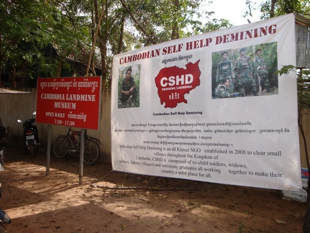 Weißes Infoplakat des Cambodian Self Help Demining mit rotem Länderkontour und Soldatenfotos vor Bäumen