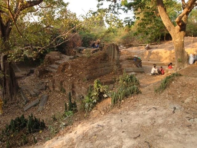Ausgrabungsgelände mit Steinmauern, Pflanzen und sitzenden Personen unter Bäumen in Angkor, Kambodscha