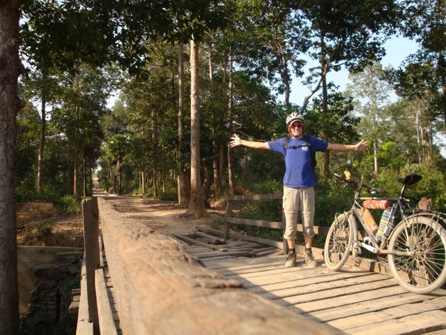 Radfahrer in blauem Shirt auf Holzsteg zwischen Bäumen, Fahrrad im Hintergrund, sonnige Waldlandschaft