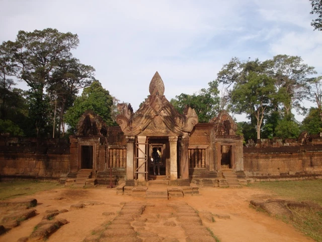 Historische Sandstein-Tempelarchitektur mit Eingangsbogen, umgeben von Bäumen, rötlich-brauner Steinboden