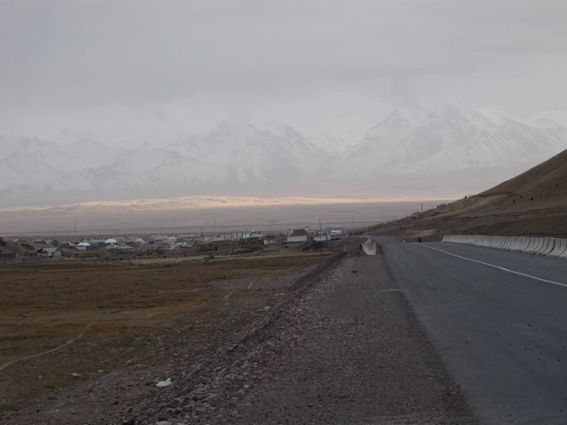 Asphaltierte Straße führt an kahler Landschaft vorbei, im Hintergrund Berge und kleine Häuser, grauer Himmel