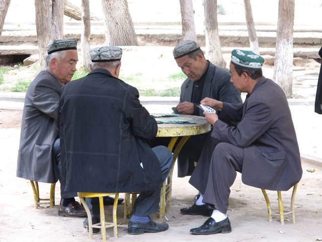 Vier ältere Männer in traditioneller Kleidung sitzen an einem gelben Tisch und spielen Karten, umgeben von Bäumen