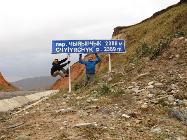 Zwei Personen posieren an einem blauen Straßenschild für einen Pass in steiniger Berglandschaft mit 2389 Metern Höhe