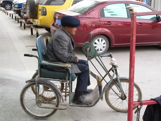 Älterer Mann in Jacke und Mütze auf modifiziertem Rollstuhl-Fahrrad mit Anhänger, parkende Autos im Hintergrund