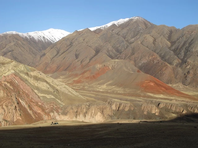 Weite Berglandschaft mit mehreren Gebirgszügen in Braun-, Rot- und Grüntönen, schneebedeckte Gipfel im Hintergrund