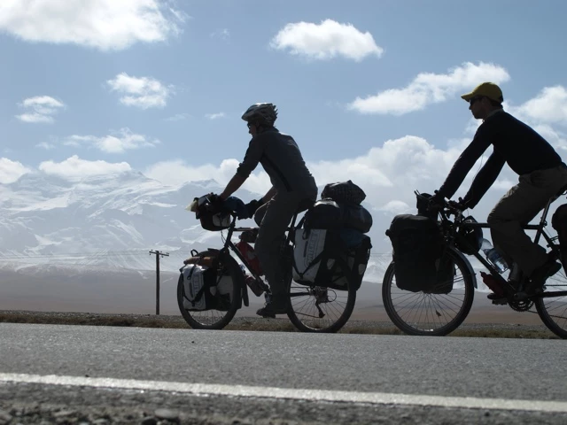 Zwei Radfahrer mit Gepäcktaschen auf Landstraße, Berge und Wolkenhimmel im Hintergrund, Silhouetten vor hellem Horizont