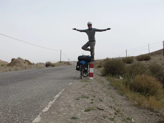Radfahrer steht auf Fahrrad mit ausgebreiteten Armen, neben einer einsamen Straße in karger Landschaft